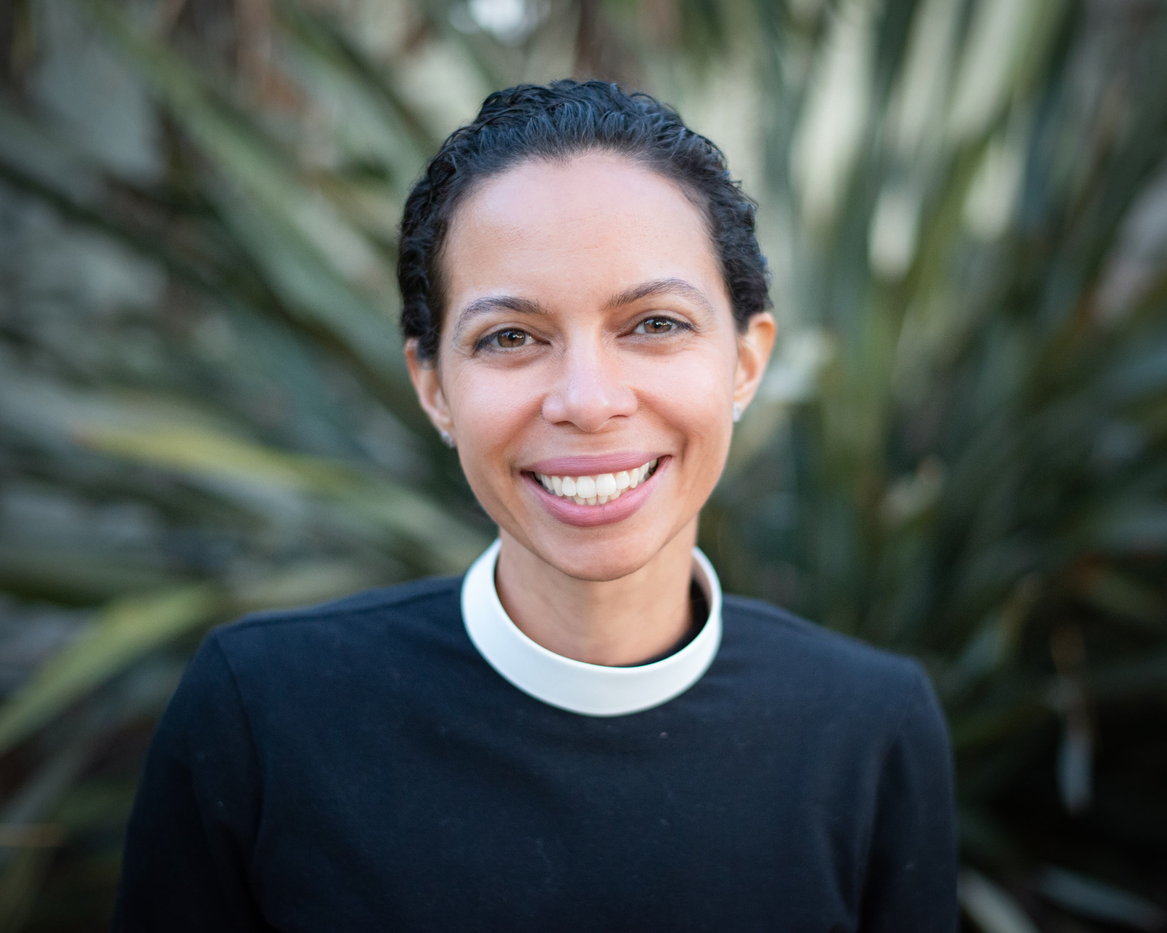 Rev. Sally Steele standing outdoors in a forested natural setting wearing a vest, looking confidently at the camera